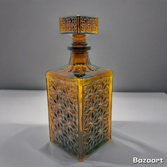 A square-shaped vintage decanter with a dark amber brown color and an all-over geometric pattern, accompanied by a matching stopper. The decanter is empty and displayed against a grey background.